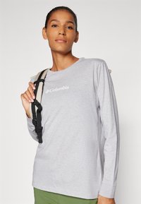 Langärmliges graues Shirt mit rundem Ausschnitt, versehen mit einem weißen Columbia-Logo, kombiniert mit einem Rucksackriemen über der Schulter.
