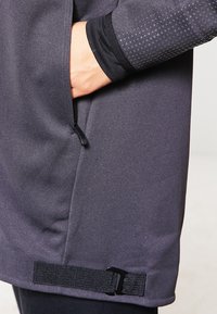 Giacca performante grigia con maniche testurizzate, tasca con zip e cinturino regolabile all'orlo. Tessuto liscio con un design aderente.