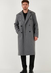 Uomo che indossa un lungo cappotto grigio doppiopetto con bottoni neri, pantaloni neri e scarpe nere, in piedi con una mano in tasca.