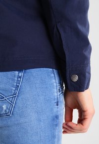 Granatowy rękaw kurtki z zapięciem na guzik, o gładkiej fakturze i starannym przeszyciu; w połączeniu z jasnoniebieskimi dżinsami.