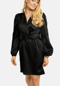 Melna, ceļgalu garuma kleita no gludas auduma ar pārsiešanas dizainu, garām pūkainām piedurknēm un izkliedētām baltām punktu rakstām.