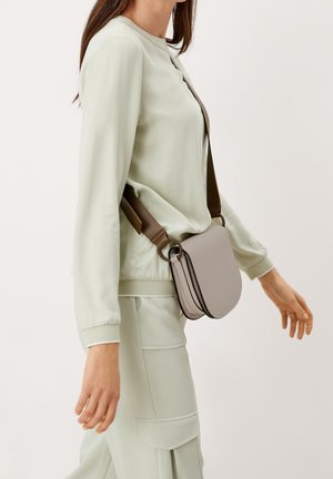 Sac bandoulière gris à forme courbée, bordure fine noire et une sangle marron. Porté avec un haut vert clair et un pantalon, créant un look doux.
