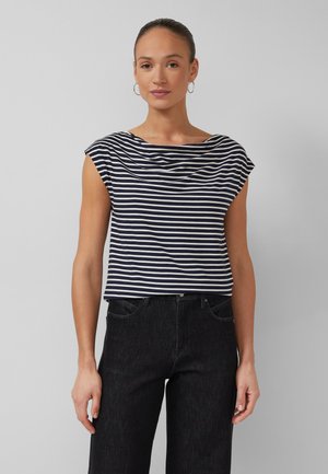 Woman with hair tied back wearing hoop earrings, a navy and white striped sleeveless top, and black high-waisted jeans against gray background.