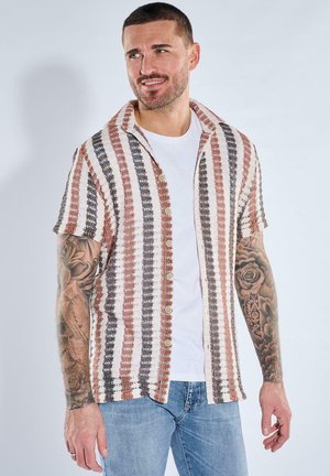 Man with short hair and tattoos wears a short-sleeve, button-up striped shirt over a white tee and blue jeans, standing against a plain background.