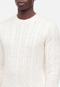 Homme portant un pull blanc en maille torsadée avec un col rond, montré des épaules à la moitié du torse sur un fond uni.