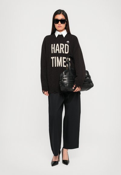 Black knit sweater with “HARD TIMES” text, white collared shirt underneath, black wide-leg pants, large black handbag, and black pointed shoes.