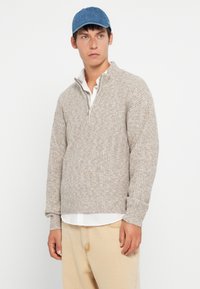 Nuori mies, jolla on päässään sininen lippis, beige ribatty vetoketjulla varustettu villapaita valkoisen paidan päällä ja vaaleanbeiget housut, seisoo tasaisen valkoisen taustan edessä.