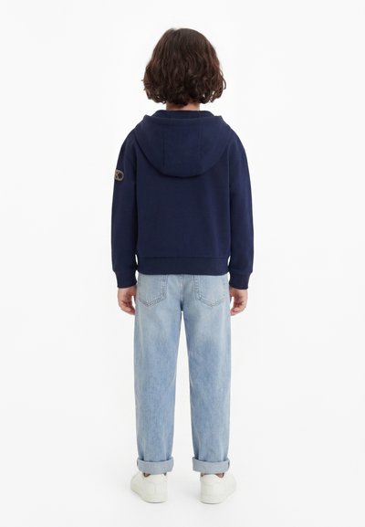 Enfant aux cheveux courts bouclés marron, portant un sweat à capuche bleu marine, un jean bleu clair retroussé et des baskets blanches, debout de dos.