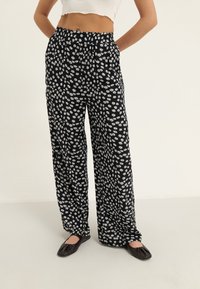 Pantalon noir à jambes larges avec un motif floral blanc, fabriqué en matière légère avec une taille élastique et une coupe décontractée.