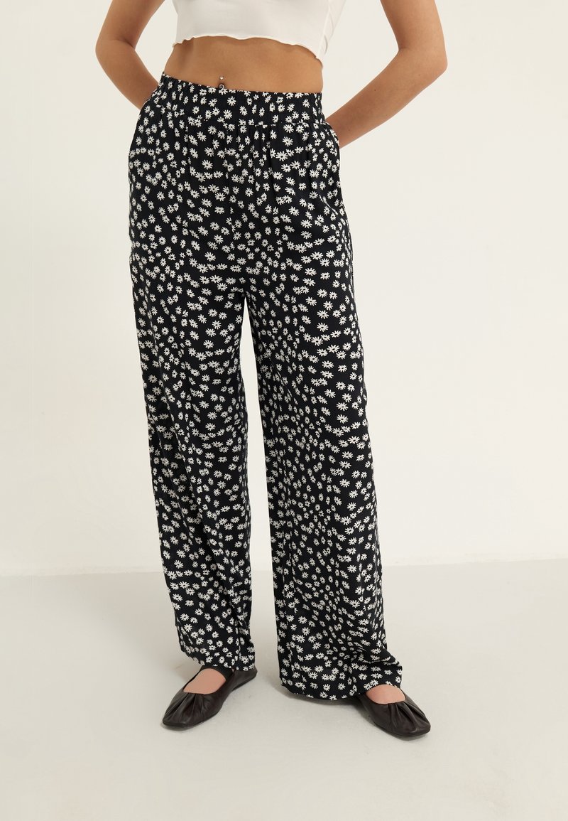 Pantalon noir à jambes larges avec un motif floral blanc, fabriqué en matière légère avec une taille élastique et une coupe décontractée.