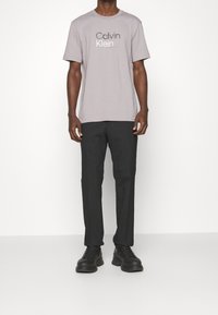 Lys grå bomulds-T-shirt med "Calvin Klein" logo, sammen med sorte bukser. Sorte kraftige snørebåndssko fuldender outfittet.