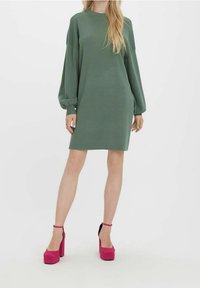 Vestito verde a maniche lunghe con una vestibilità rilassata, dotato di scollatura rotonda. Abbinato a una zeppa fucsia, creando un contrasto audace.