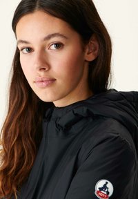 Zwarte hooded jacket met een soepele textuur, voorzien van een logopatch op de mouw en een verstelbare trekkoord rondom de capuchon.