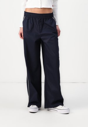 Pantalons larges bleu marine en tissu léger, dotés d'une taille élastique et de détails de piping sur les côtés. Associés à des baskets blanches.