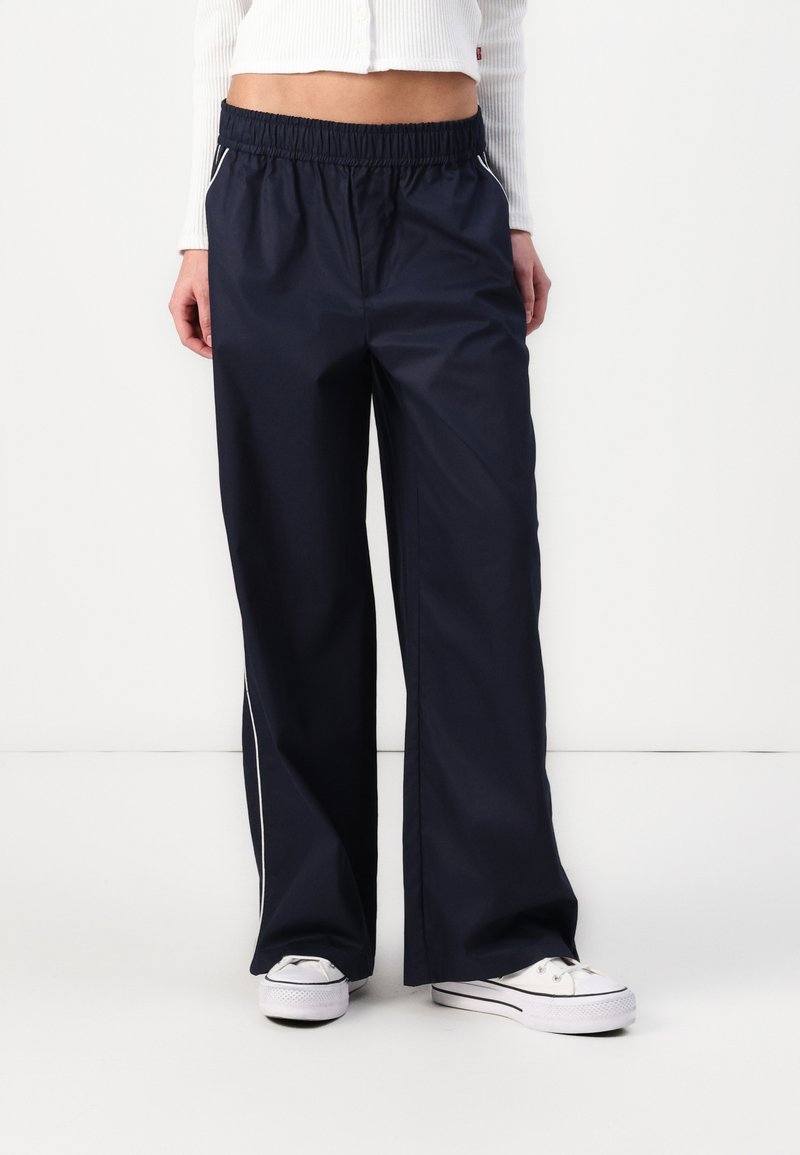 Marineblaue, weit geschnittene Hose aus leichtem Stoff, mit elastischem Bund und seitlichen Paspelierungen. Kombiniert mit weißen Sneakern.