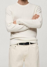 White ribbed long-sleeve sweater with a round neckline, paired with light-colored pants and a black belt. Neutral tones and textured fabric.