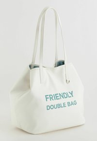 Hvid tote taske med tekstureret overflade, med to håndtag og trykt "VENLIG DOBBELT TASKE" i teal. Indvendigt er den også teal.