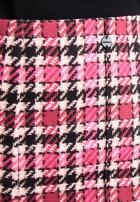 Jupe en pied-de-poule rose et noir, avec un tissu texturé, présentant une couture visible et un accent métallique de marque près de la taille.