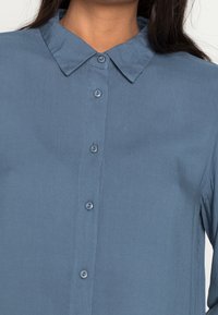Chemise bleue à boutons avec col, présentant une texture lisse et six boutons sur le devant. Design simple sans motifs ni accents.