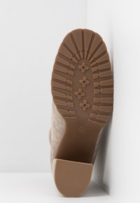 Semelle de botte en suède beige avec des motifs de bande de roulement profonds, des grips en forme de fleur, et un talon texturé, placée contre un mur d'angle blanc.