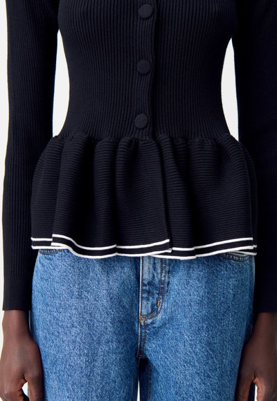 Cardigan nero a coste con bottoni coperti in tessuto e orlo peplum rifinito in bianco abbinato a jeans blu medio su una persona di carnagione scura.