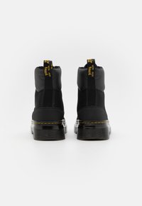 Paire de bottines noires en cuir et tissu avec semelles épaisses et coutures jaunes, vues de dos sur fond blanc.