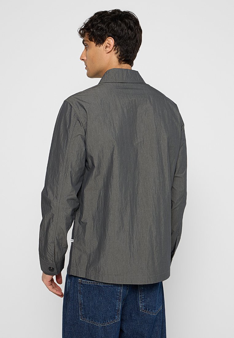 Szara, lekka kurtka o teksturowanym wykończeniu, z zapięciem na guziki na mankietach i prostym kroju, noszona na niebieskich dżinsach. Widok z tyłu.