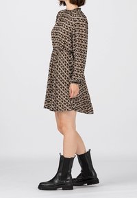 Hnědé šaty s vzorem, vysokým výstřihem a dlouhými rukávy, se zúženým pasem a rozšířenou sukni, doplněné černými kotníkovými botami.