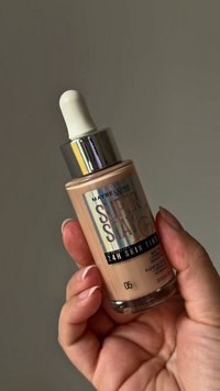 Ręka trzymająca butelkę podkładu Maybelline Super Stay 24H Skin Tint z kroplomierzem, odcień numer 05, na jednolitym tle.