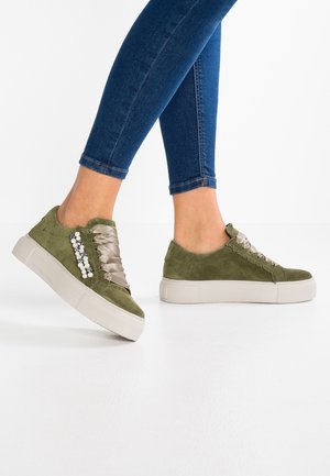 Kvinde iført olivengrønne ruskindsplatformsneakers med satinbånd og perledekorationer, stylet med tætsiddende blå jeans mod hvid baggrund.