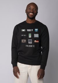 Black sweatshirt featuring retro gaming console graphics in various colors. Text below reads "I'M LOVIN' IT." Smooth fabric, casual fit.