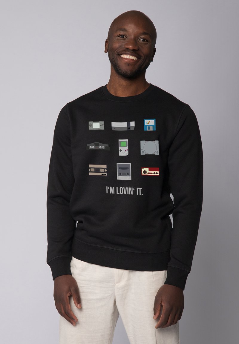 Black sweatshirt featuring retro gaming console graphics in various colors. Text below reads "I'M LOVIN' IT." Smooth fabric, casual fit.