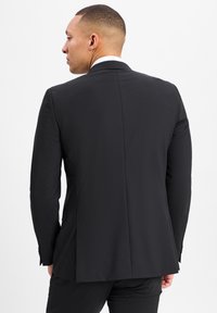 Zwarte blazer met een getailleerde pasvorm, gestructureerde schouders, een enkele split aan de achterkant en een subtiele textuur in de stof. Knopdetail bij de manchetten.