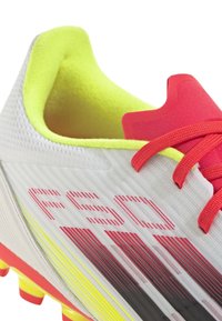 Jalkapallokengät, joissa on valkoinen ja kirkas keltainen päällinen, punaiset nauhat ja raidallinen kuvio. Kevyt muotoilu teksturoitua materiaalia ja rohkeaa brändäystä.