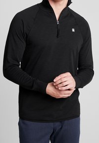 Svart långärmad pullover med kvartdragkedja, med en slät yta, raglanärmar och en liten logotyp på bröstet.