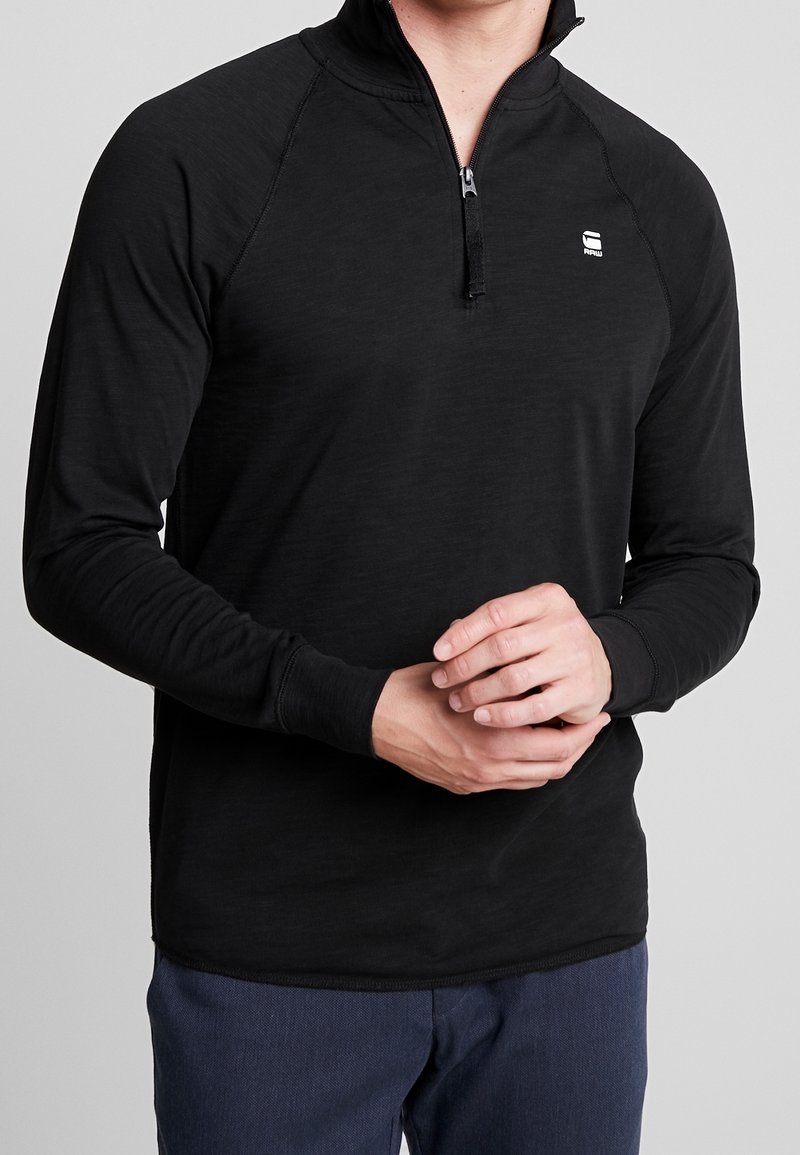 Svart långärmad pullover med kvartdragkedja, med en slät yta, raglanärmar och en liten logotyp på bröstet.