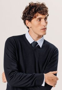 Pull en v marine en tissu doux sur une chemise à rayures bleues claires, avec une coupe slim et des poignets côtelés, associé à une cravate sombre.