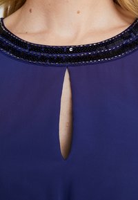 Fialové šaty s kulatým výstřihem zdobeným černými flitry a malým klíčovým otvorem v přední části. Hladká textilie.