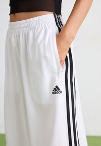 Valkoiset urheilushortsit, joissa on kiristysnauha vyötäröllä, mustat sivuraitat ja Adidas-logo. Materiaali näyttää olevan sileää ja kevyttä.