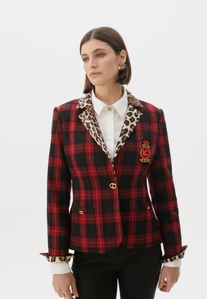 Modella che indossa un blazer a quadri rosso e nero con colletto maculato e una camicia bianca, rivolta leggermente verso destra.
