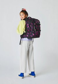 Sac à dos noir avec des motifs floraux violets et des accents roses. Le modèle porte un pull vert clair et un pantalon blanc, avec des chaussures bleues.