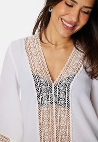 La blusa bianca presenta un intricato dettaglio in pizzo floreale nero lungo il collo e la parte anteriore. Tessuto morbido e leggero con maniche a campana.