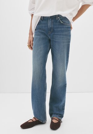 Persoon draagt blauw spijkerbroek met rechte pijpen, wit shirt met lange mouwen en donkerbruine platte schoenen met enkelbandjes, staand tegen een witte achtergrond.