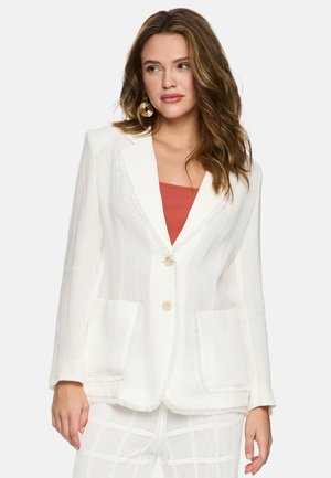 Frau mit welligem braunem Haar, die goldene kreisförmige Ohrringe, einen weißen strukturierten Blazer über einem rostfarbenen Oberteil und weiß karierte Hosen trägt.