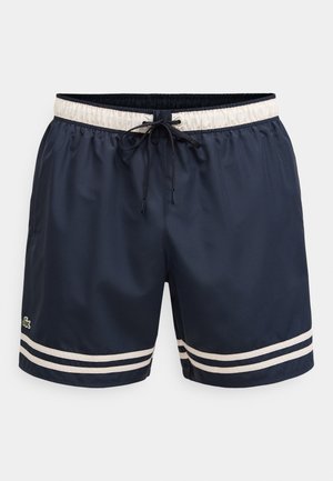 Tengerészkék fürdőnadrág fehér elasztikus derékpánttal, fekete húzózsinórral, két fehér csíkkal mindkét lábon és egy kis zöld krokodil logóval a bal lábon.