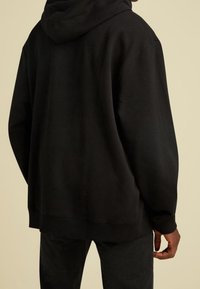 Persona che indossa un ampio hoodie nero e pantaloni neri, in piedi con la schiena rivolta verso la macchina fotografica contro uno sfondo beige uniforme.