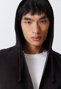 Joven con cabello corto y oscuro que lleva puesto un hoodie negro con cremallera sobre una camiseta blanca, mirando directamente a la cámara con una expresión neutral.