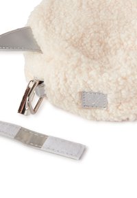 Kulatá taška vyrobená z měkké, nadýchané světle béžové látky se stříbrným popruhem a kováním, se malou šedou etiketou přišitou na přední straně.