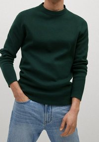 Dunkelgrüner Strickpullover mit rundem Ausschnitt, schmaler Passform, gerippten Bündchen und strukturiertem Stoff, kombiniert mit hellblauen Jeans.