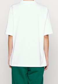 Valkoinen ylisuuri puuvillapaita rentoon istuvuuteen, lyhyet hihat ja pyöreä kaula-aukko, esitelty takaa neutraalia taustaa vasten.